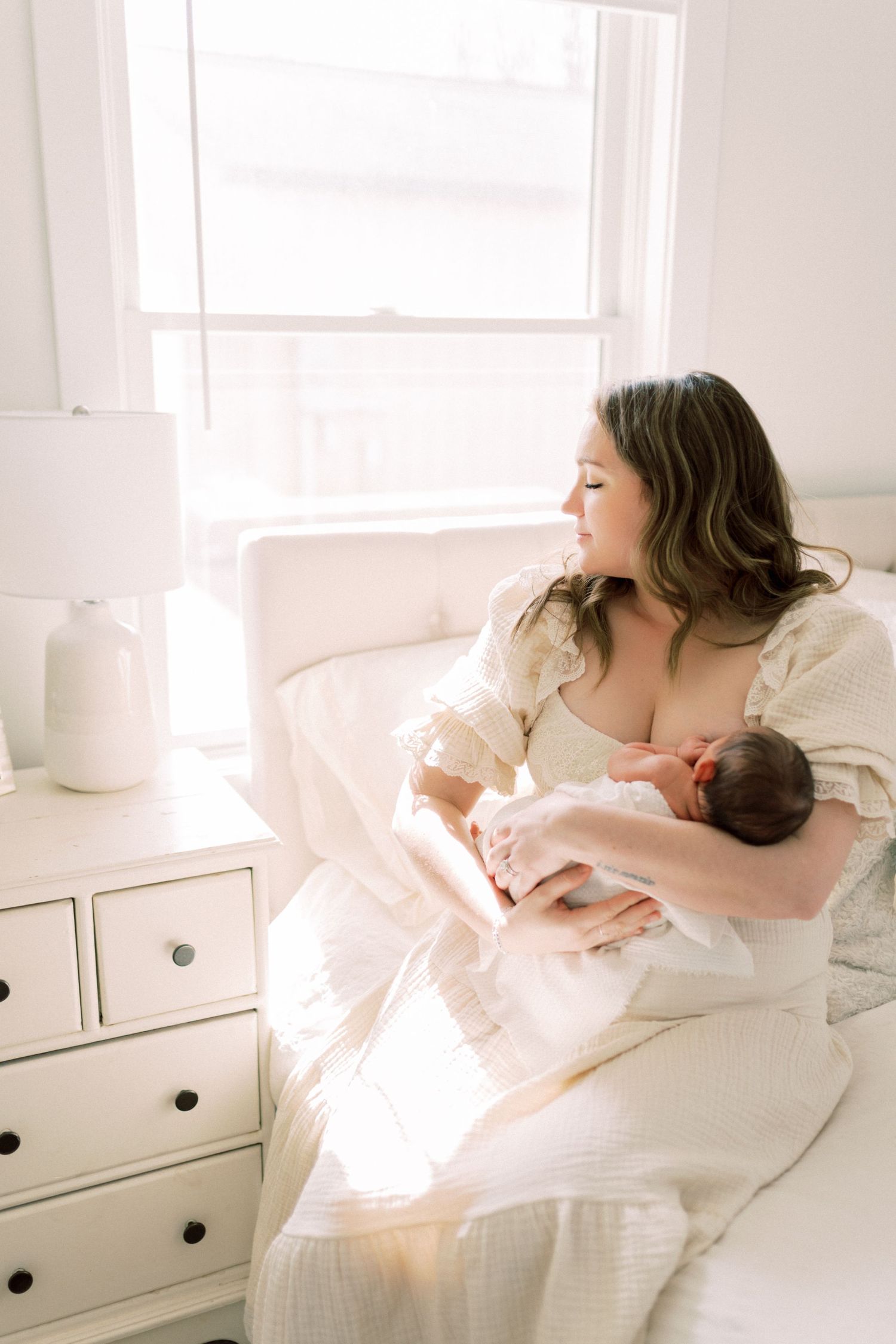 A serene moment captured in a bright white bedroom as mother breastfeeds her newborn baby next to a white dresser.