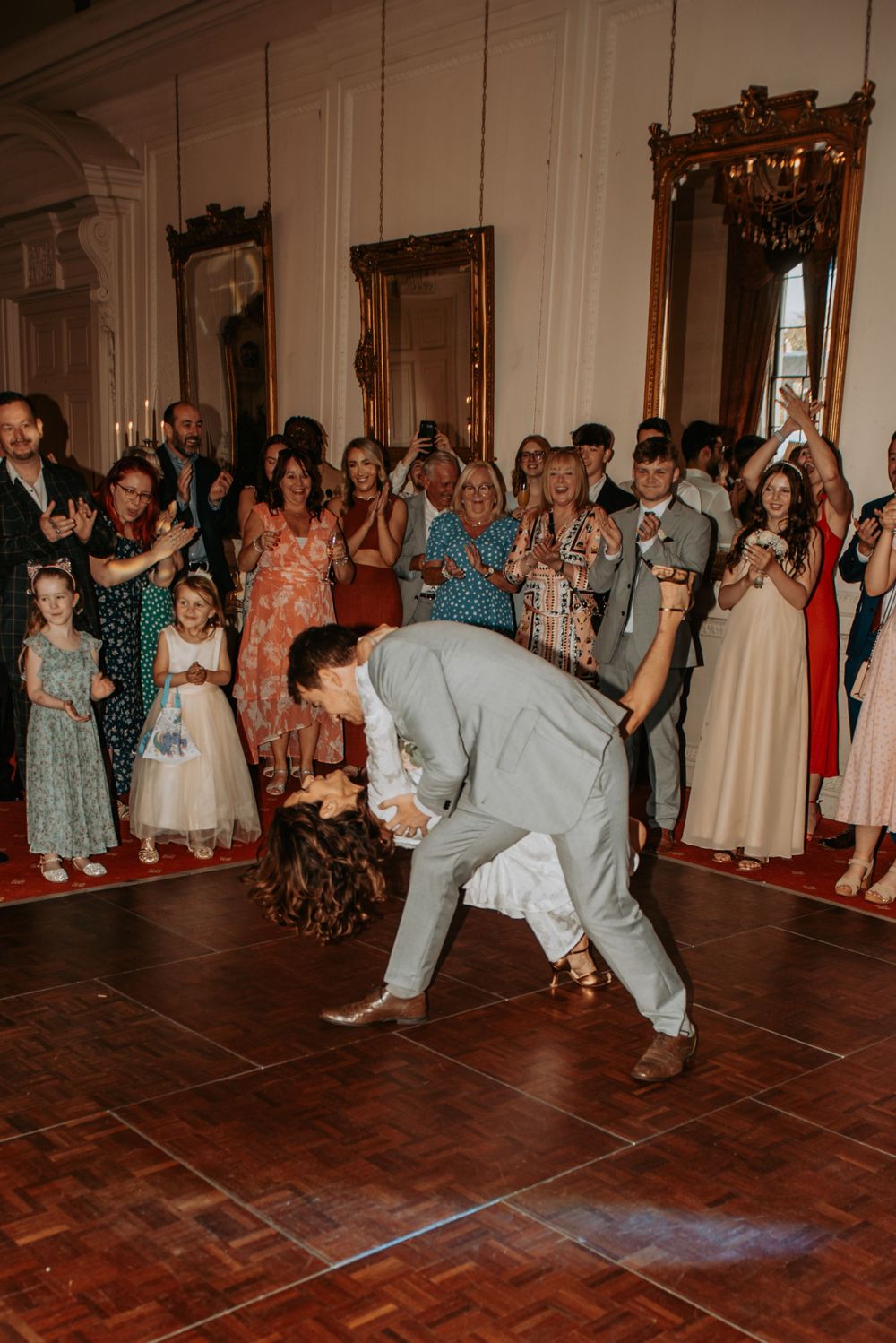 A wedding guest performs a dance move on the hardwood floor at a reception while onlookers gather around.
