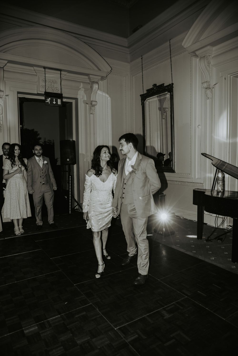 A couple walks together on a dance floor in a vintage-style black and white photograph at an elegant indoor venue.