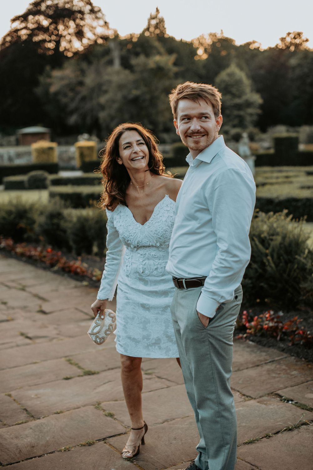 A couple in light-colored attire poses together at sunset in a garden with stone pathways and flower beds.