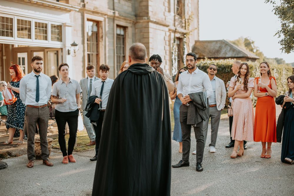 A group of wedding guests gather outdoors around someone wearing a black cape during a golden hour ceremony.