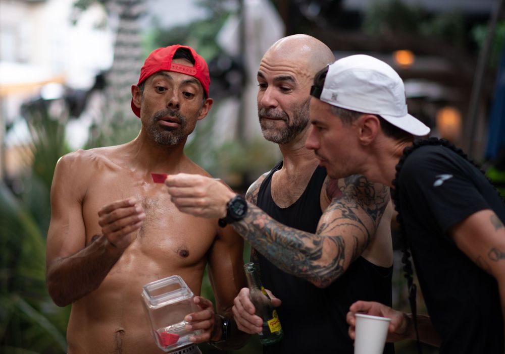 Three shirtless men having an animated discussion outdoors while holding drinks.