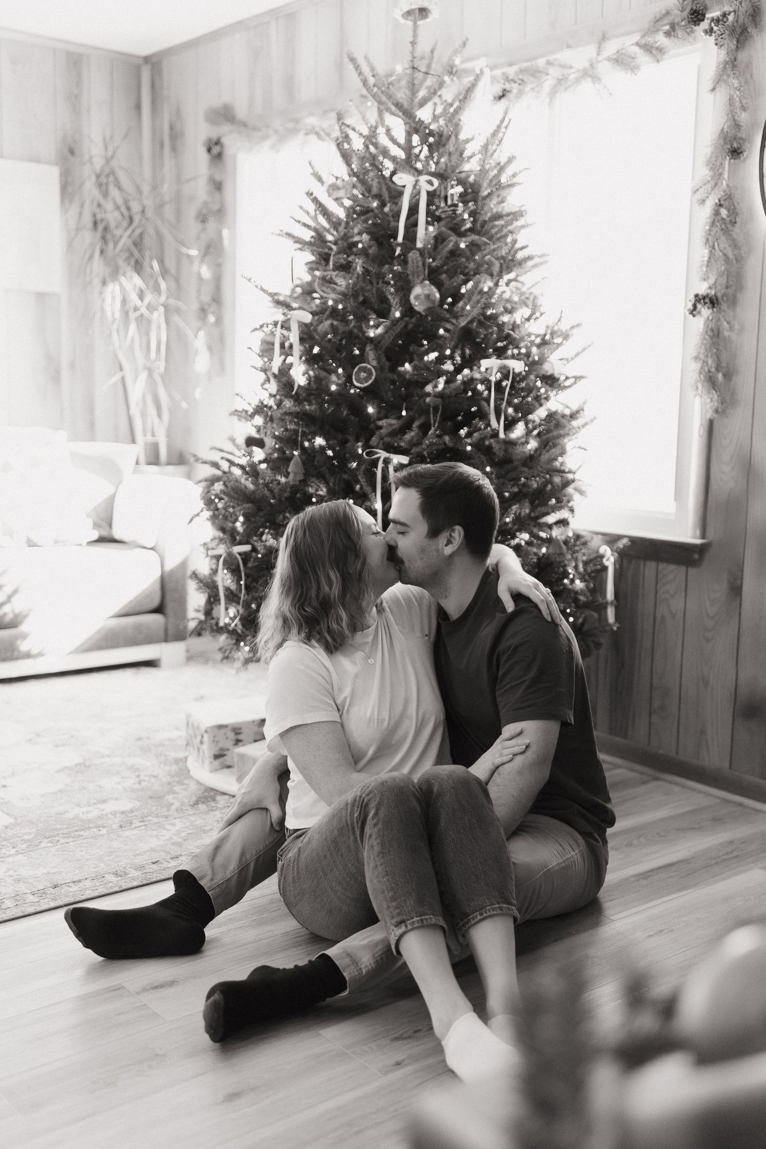 A couple sits together on the floor in front of a decorated Christmas tree.