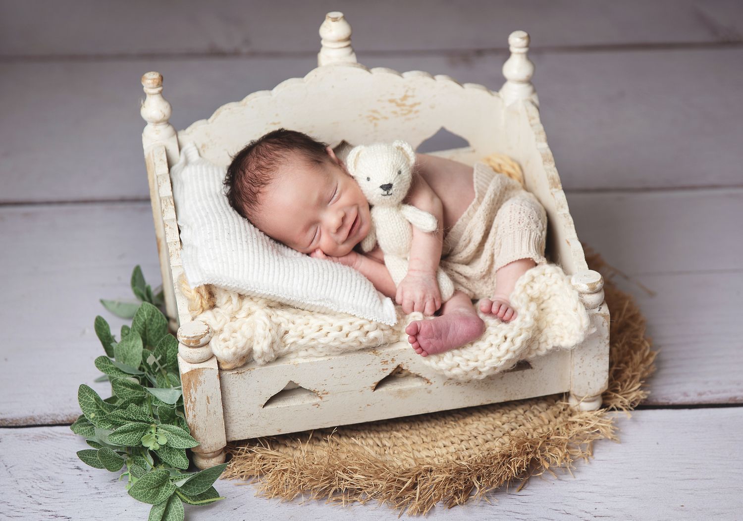 Newborn baby sleeping peacefully on rustic white wooden bed surrounded by soft cream blankets and foliage. During a photoshoot in Sidcup