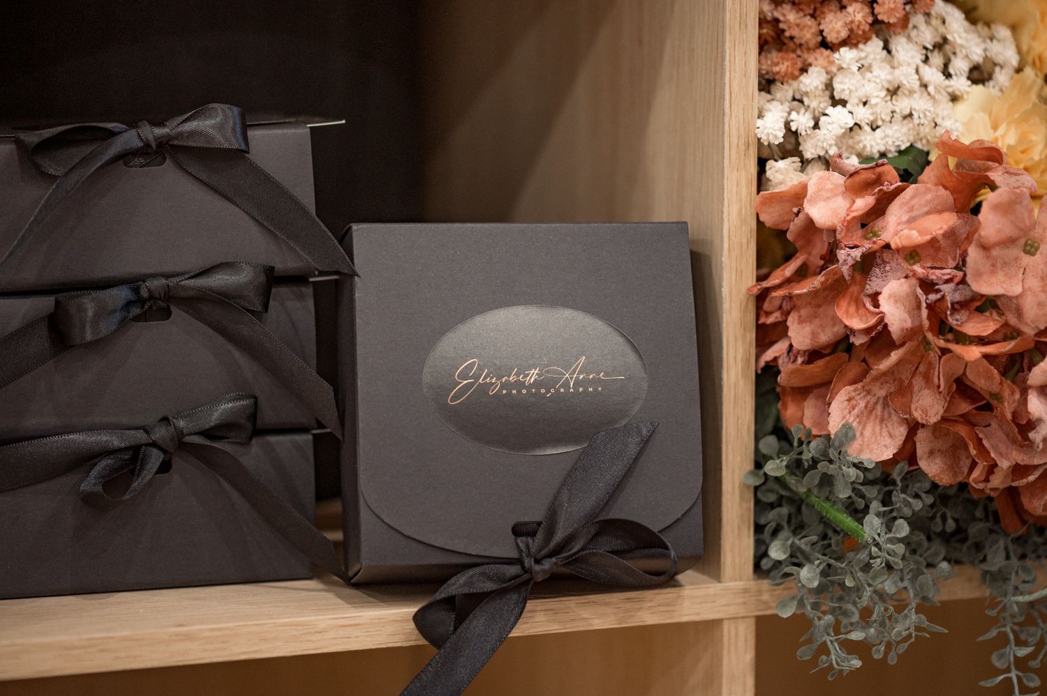 Elegant black gift boxes with grey bows arranged on a shelf next to pink floral decorations.