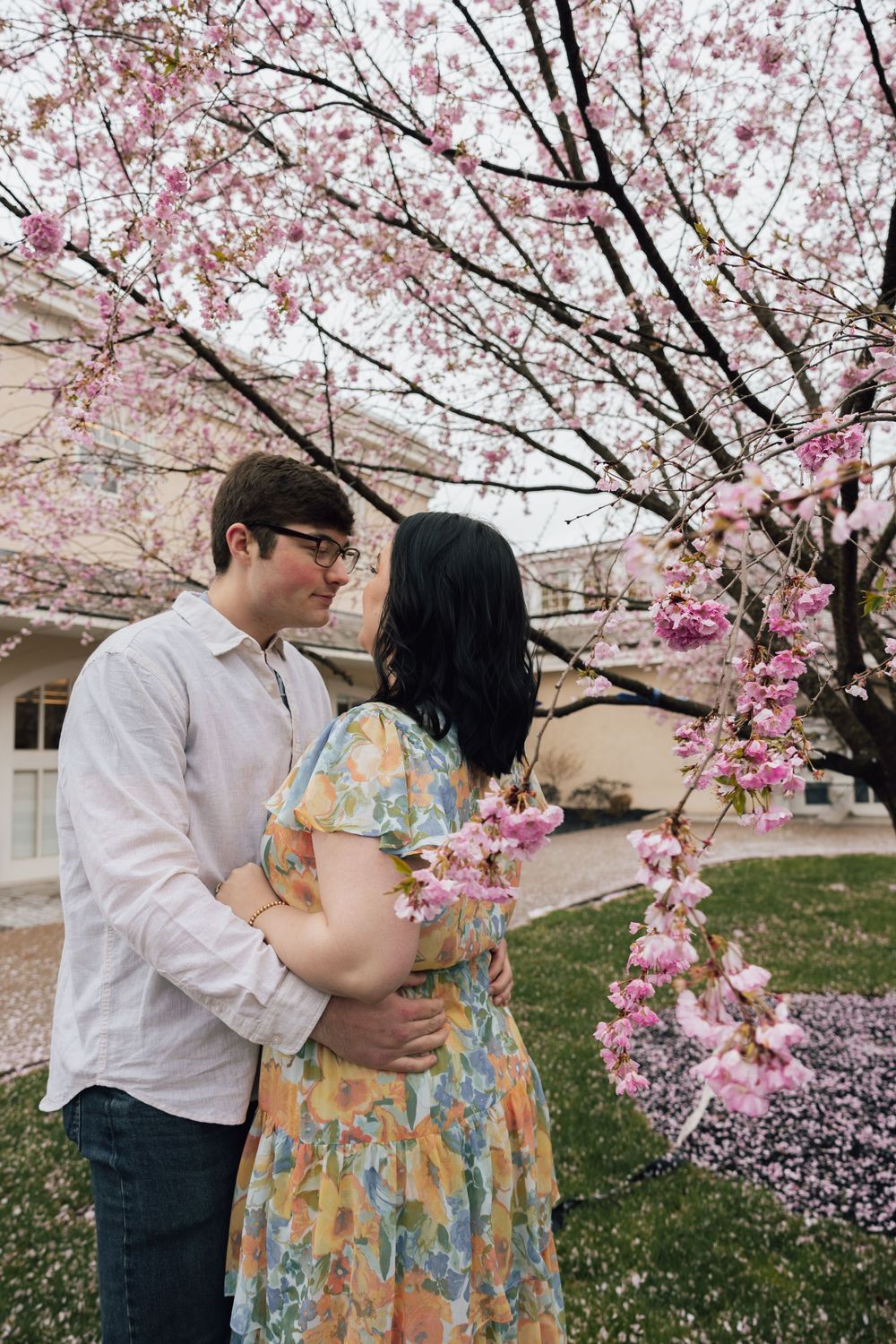 A couple embraces under blooming pink cherry blossom trees on a spring day near a building.