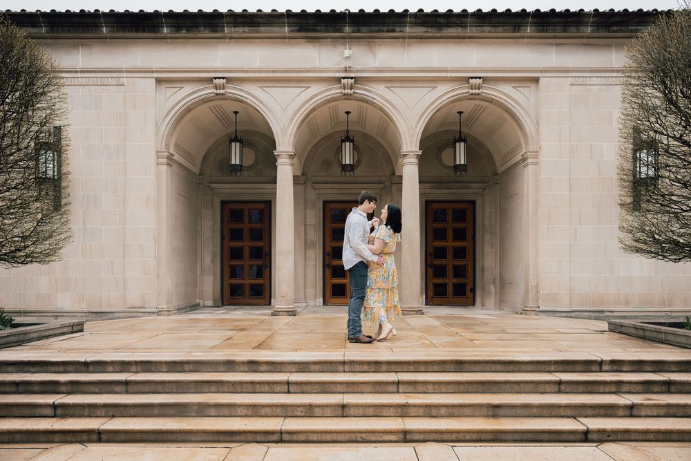 A couple embraces on the stone steps of a grand building with arched columns and wooden doors.