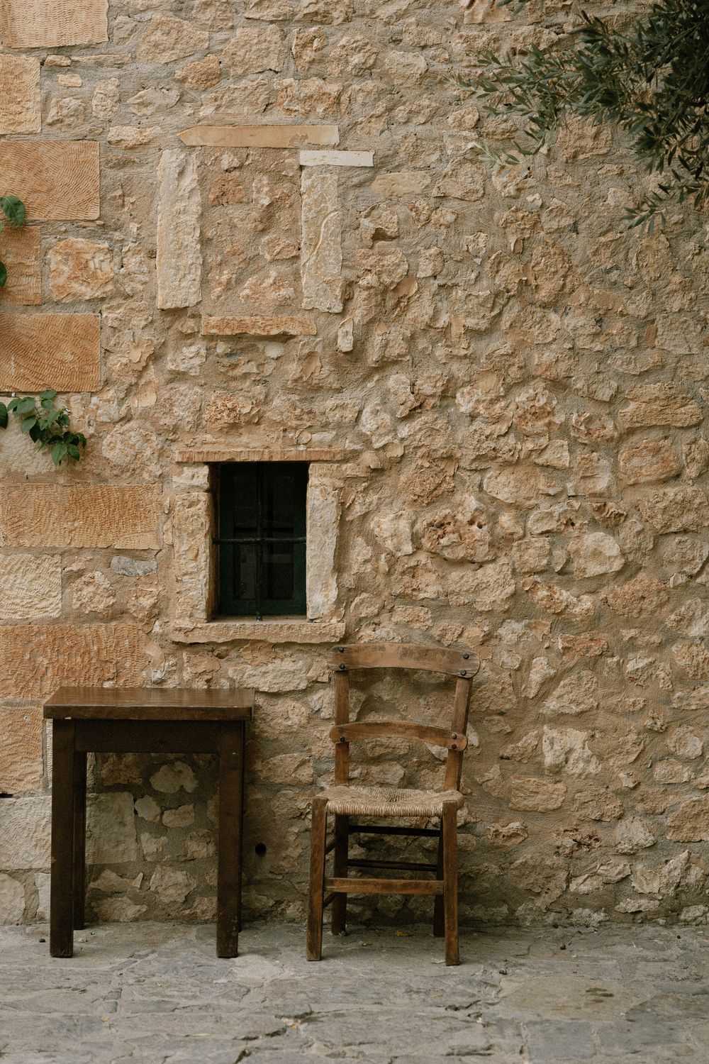 Rustic stone wall with small window, wooden chair and table creating a Mediterranean architectural scene.