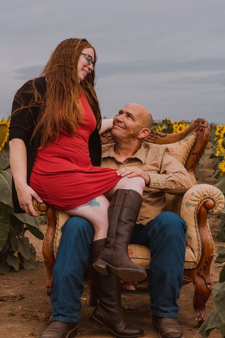 A couple sits together on an antique chair in a sunflower field, with her wearing a red dress and brown boots.