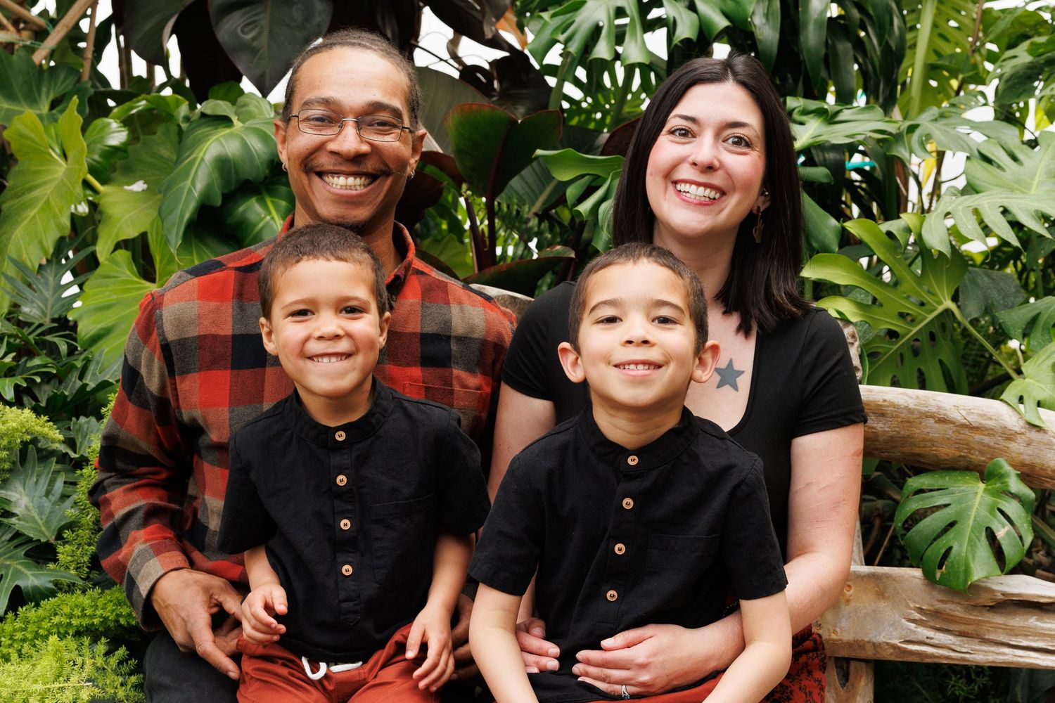 A happy family sits together in front of lush tropical foliage wearing coordinated casual outfits.