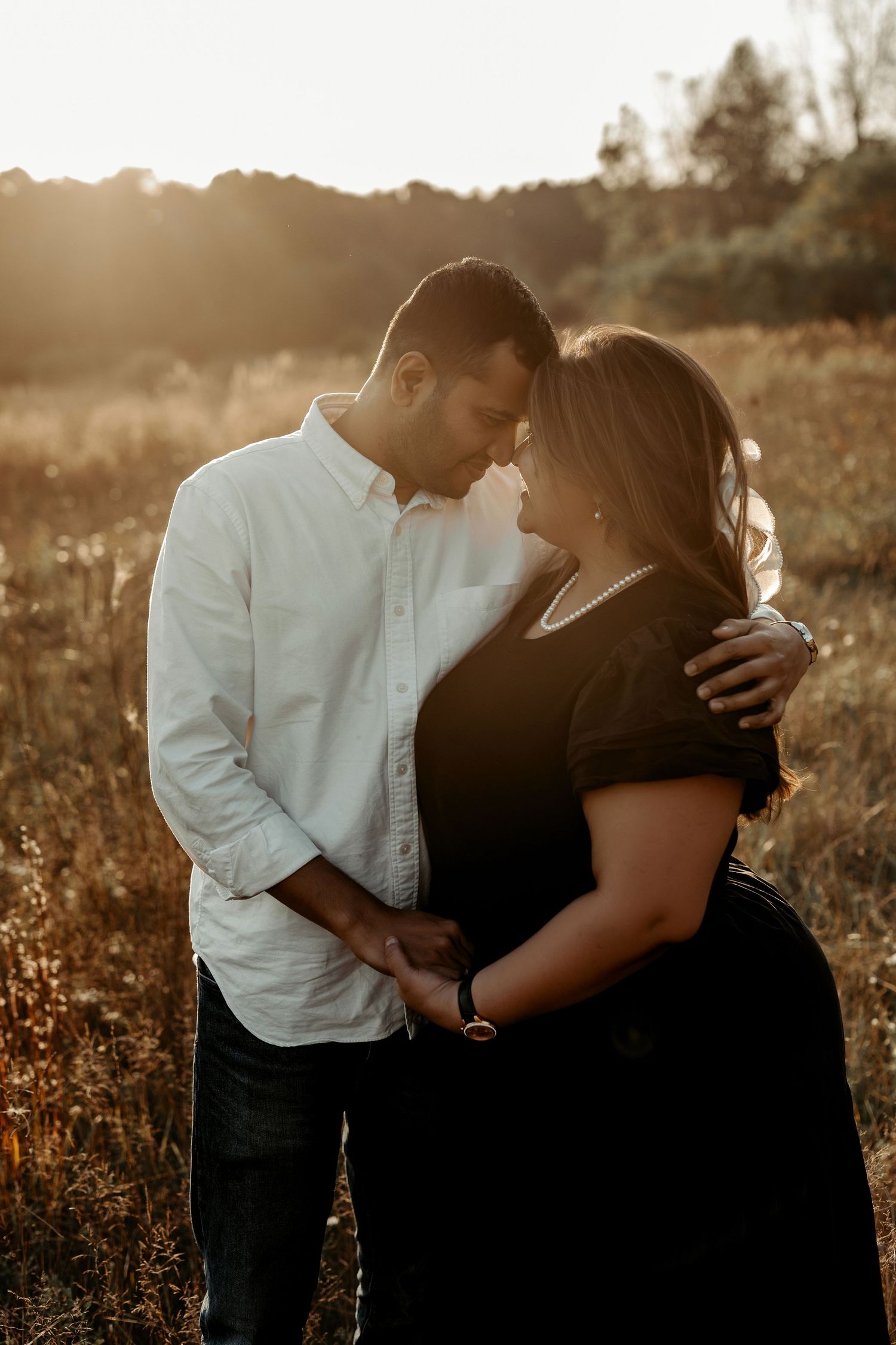 A couple shares an intimate moment in a sunlit field during golden hour as the sunsets behind them.