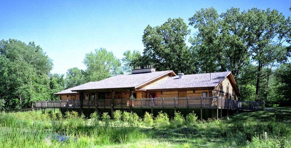 Modern ranch-style home with wooden deck surrounded by lush green trees and natural landscaping in rural setting.