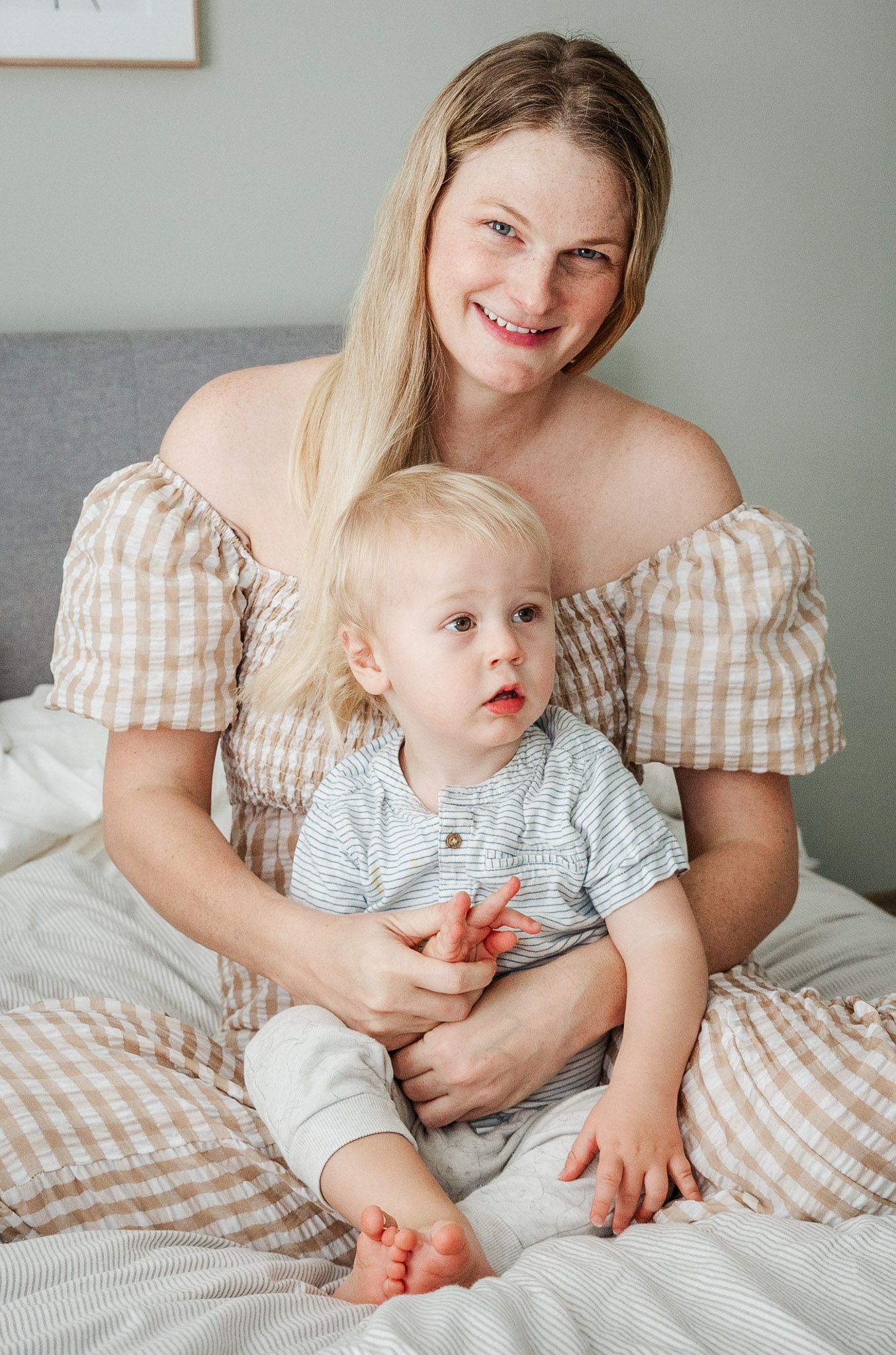 A mother smiles and looks to camera as she sits on the bed with her young son. Bristol family photos by Little Bodhi Photography