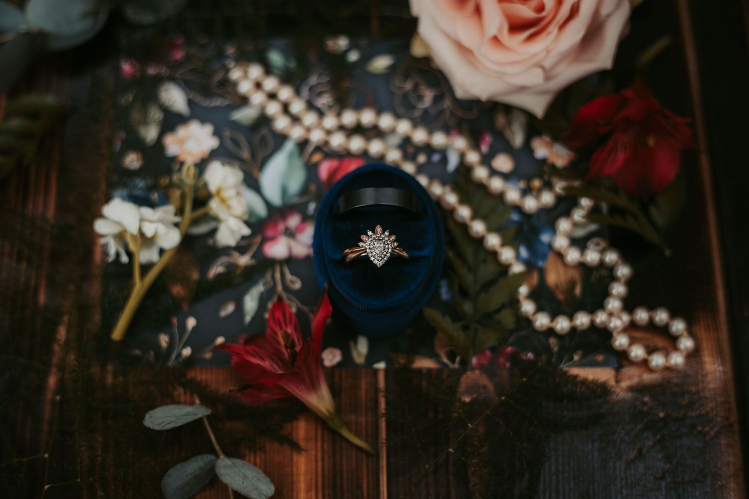 Vintage-style floral arrangement with roses, pearls, and a brooch on dark wood surface in moody lighting.