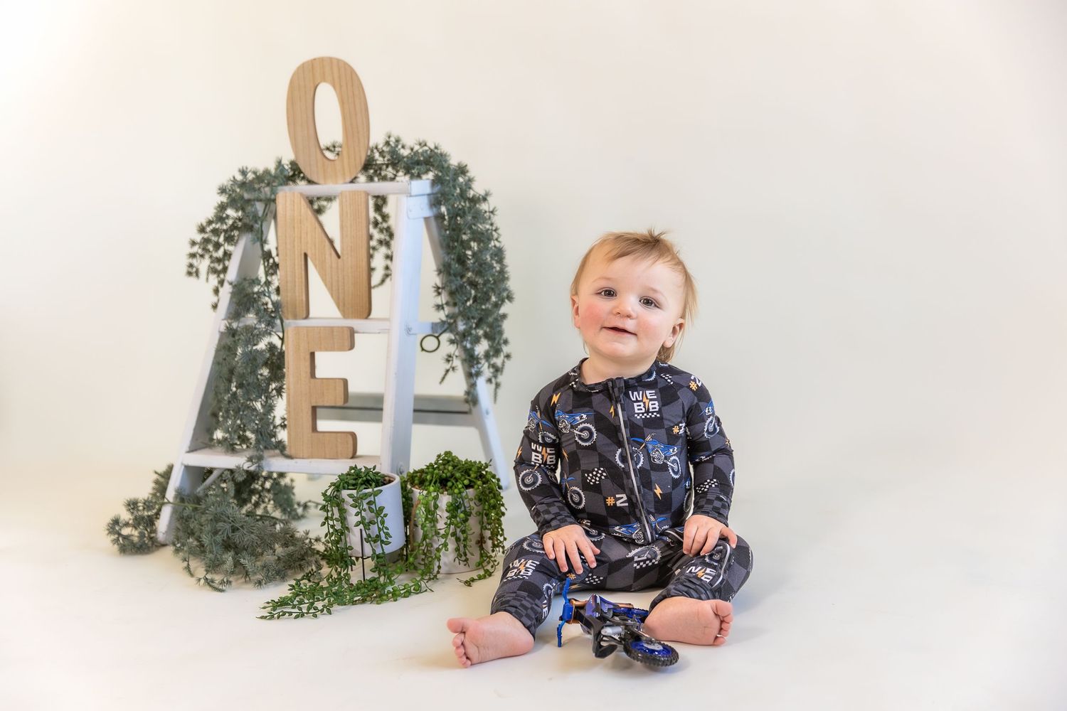 Small child in navy star print pajamas sitting next to a decorative number one display with greenery against white backdrop.