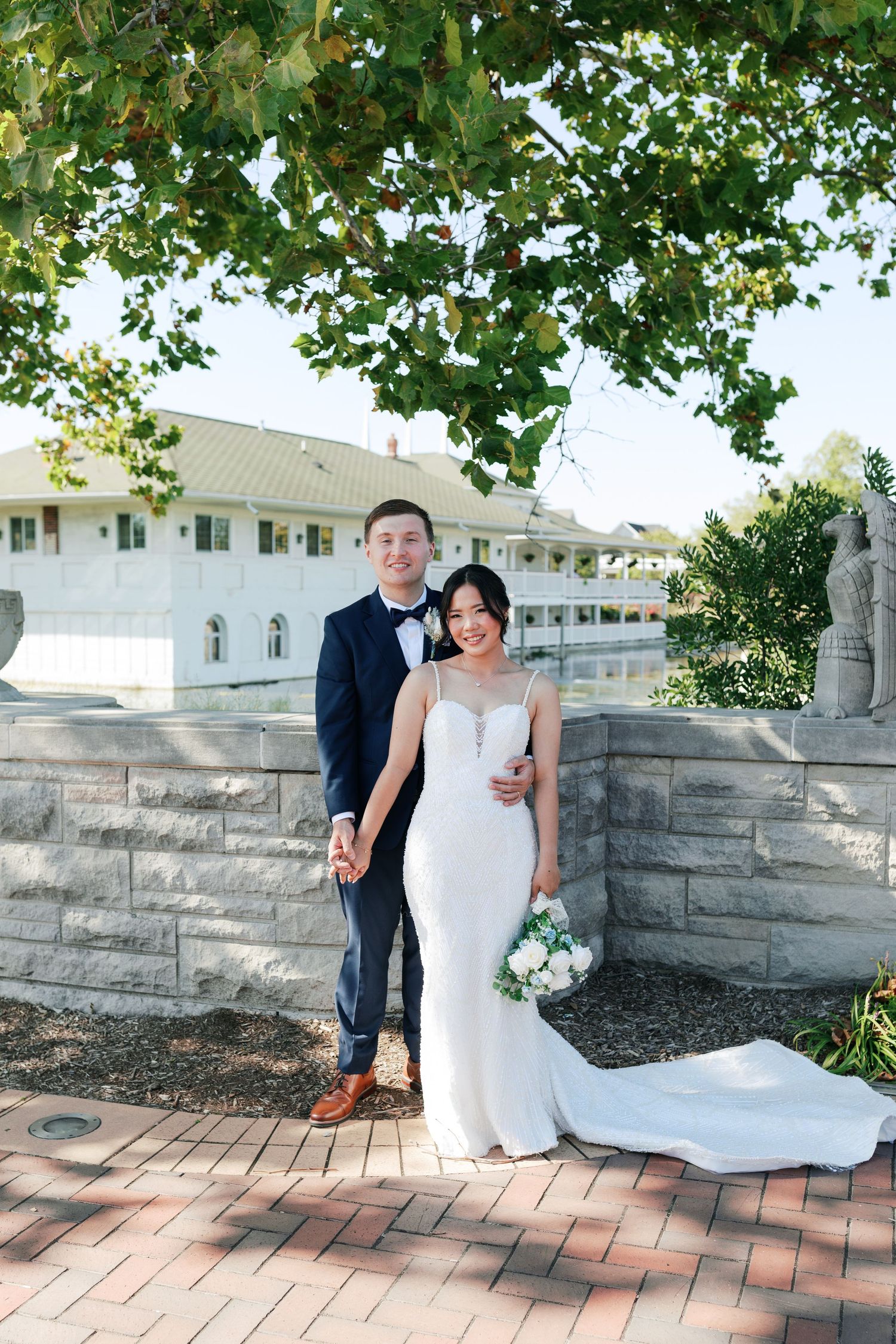 Newlyweds posing in front of historic white building with stone wall and brick pathway.