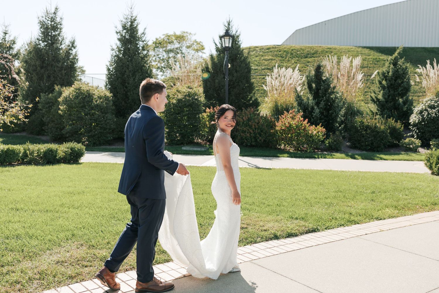 A couple walks together on a garden pathway surrounded by evergreen trees on a sunny day.