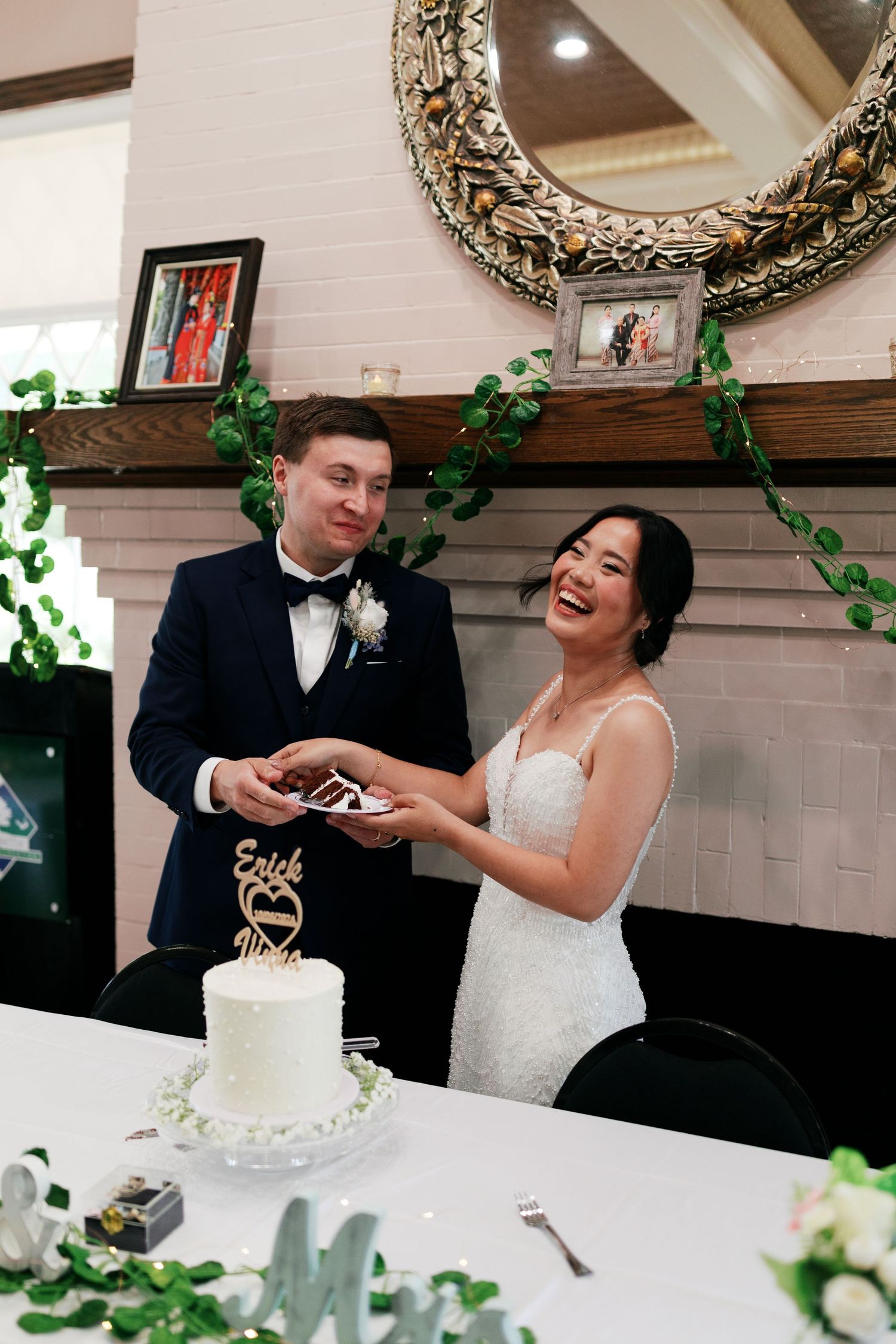 Newlyweds share a laugh while cutting their wedding cake beneath a decorative mirror.