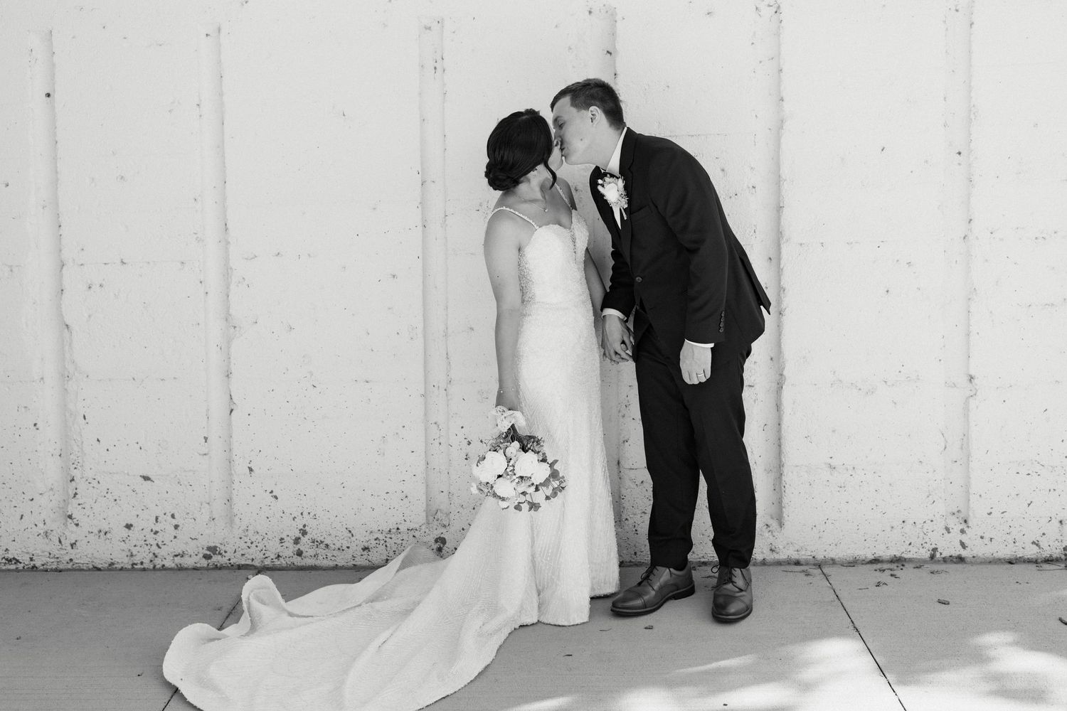 A romantic black and white portrait of newlyweds standing against a white wall.
