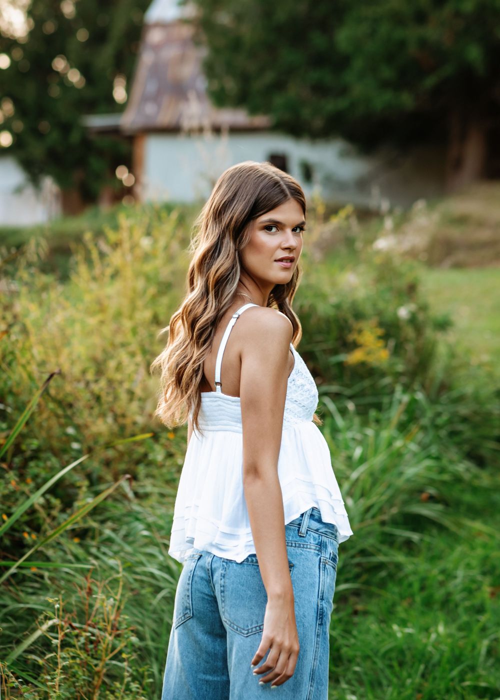 Woman in white top and jeans poses in front of lush green garden setting.