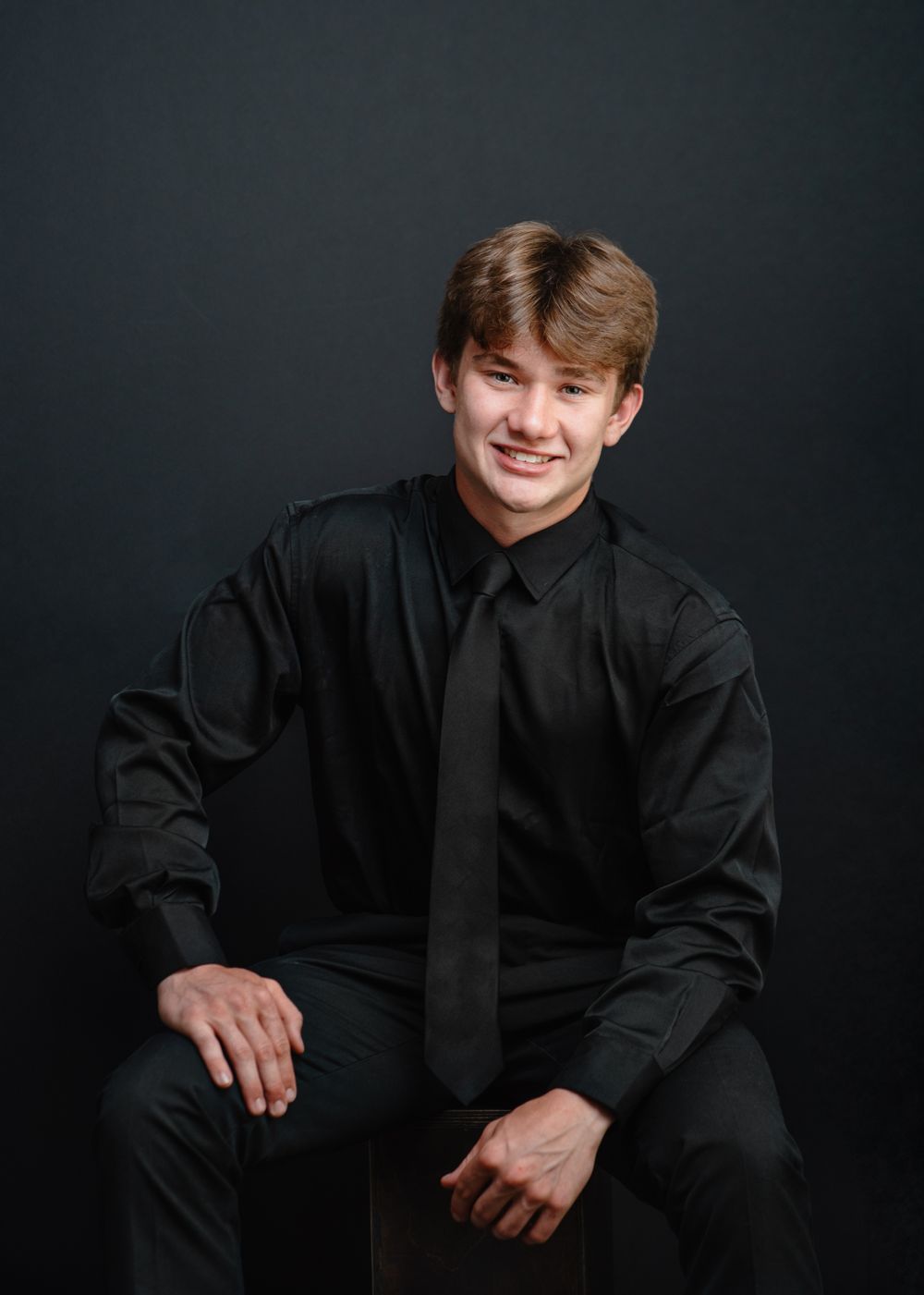 Professional studio portrait of a young person in formal black attire with black tie against dark background.