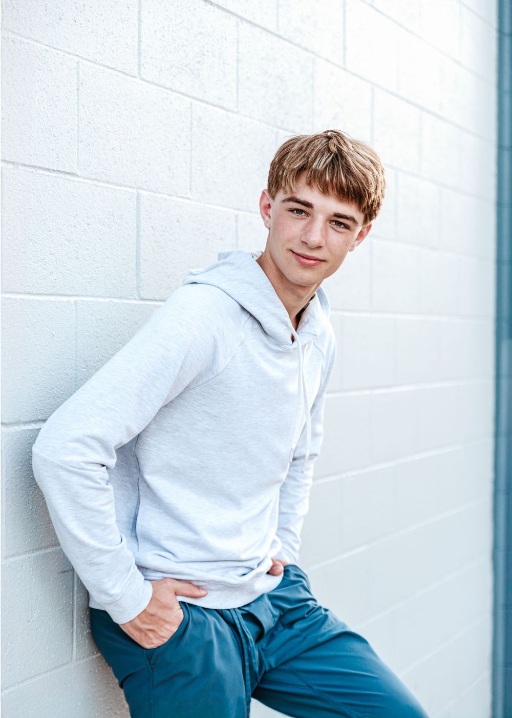 Casual portrait of person in light gray hoodie leaning against white wall.