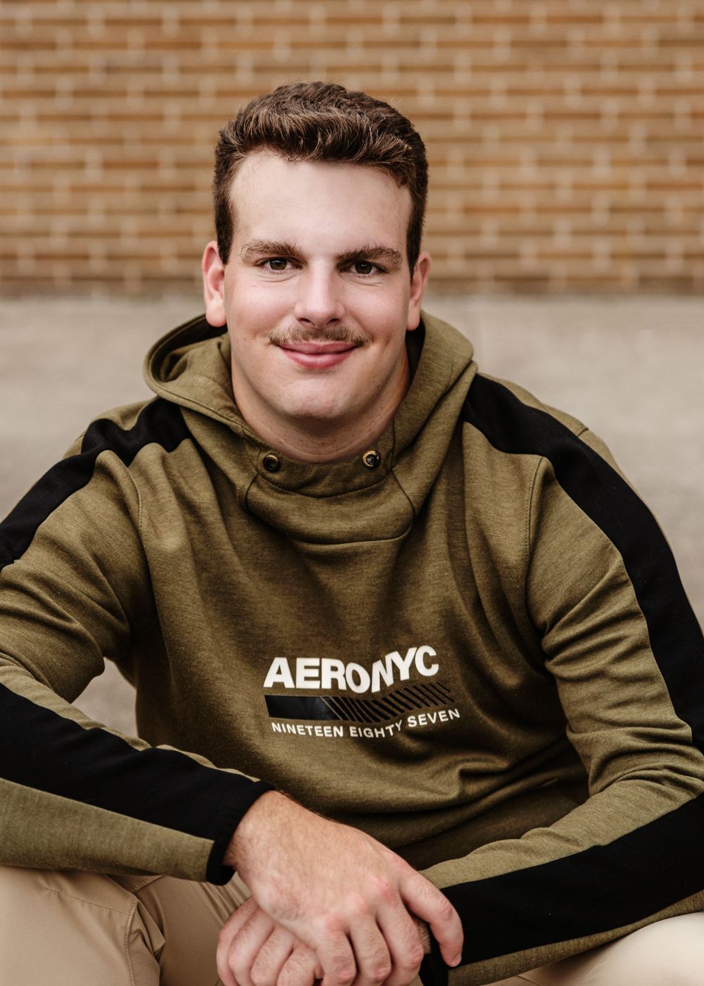 Portrait of person wearing olive green Aero NYC hoodie against brick wall background.