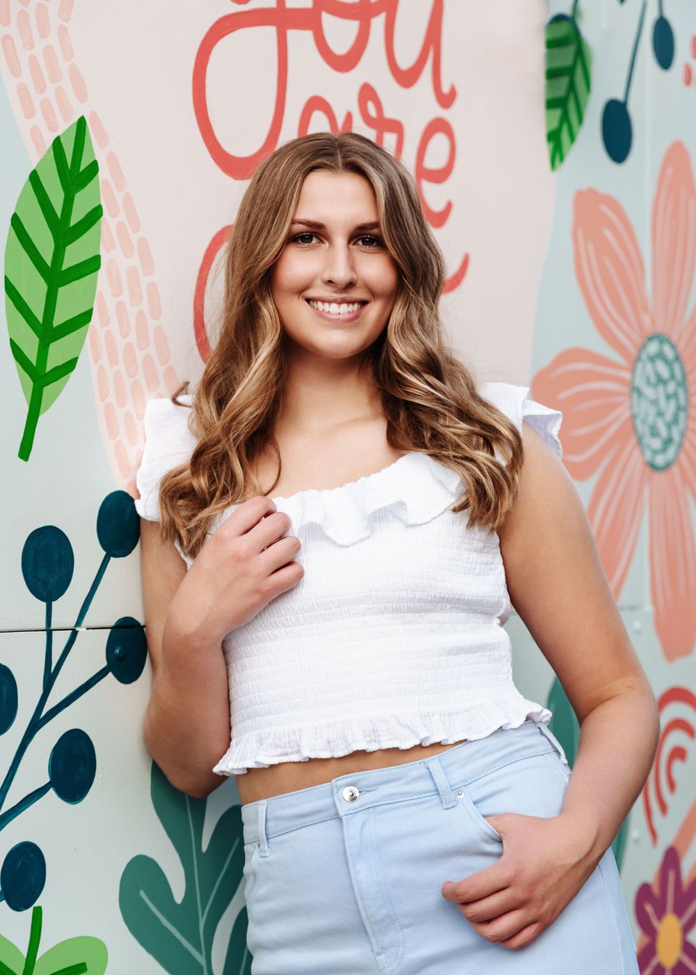 Portrait against floral mural wall wearing white ruffle crop top and light jeans.