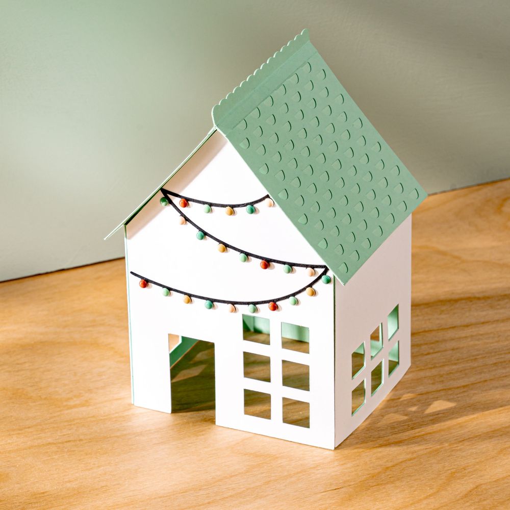 Small decorative white house model with green roof and string lights displays festive holiday charm.