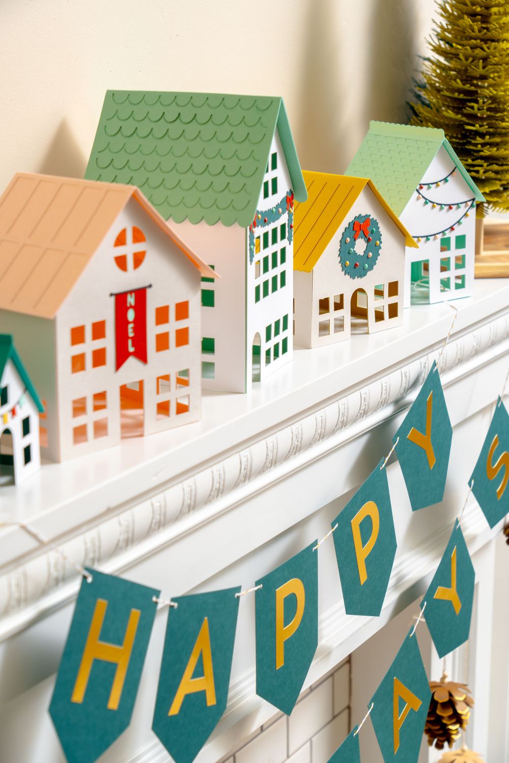 Row of miniature paper houses decorated for holidays with 'HAPPY' banner below.