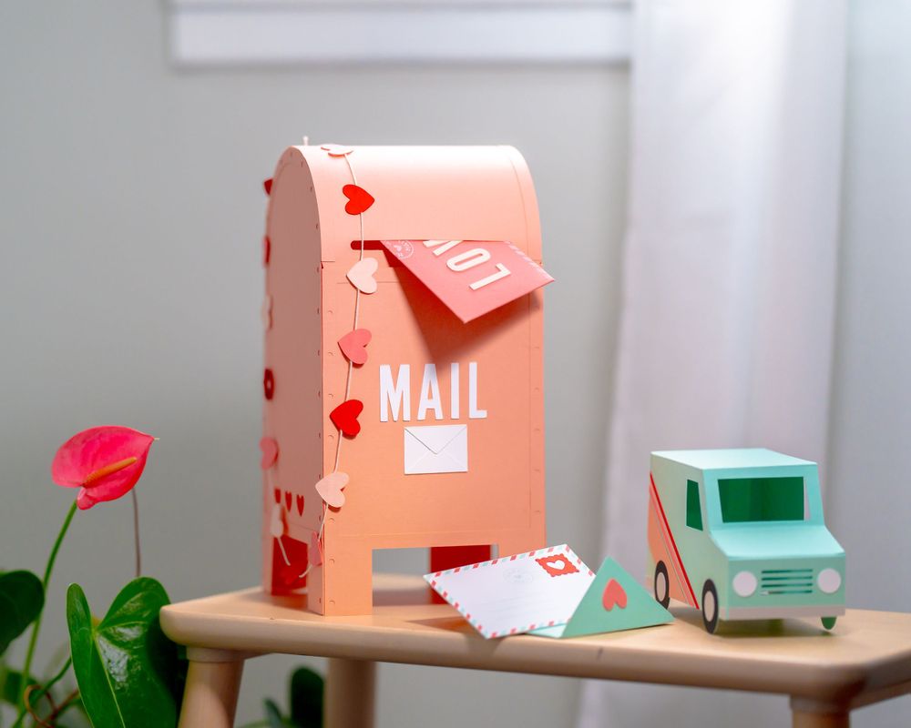 Pink decorative mailbox with heart patterns and tiny mail truck on wooden surface.