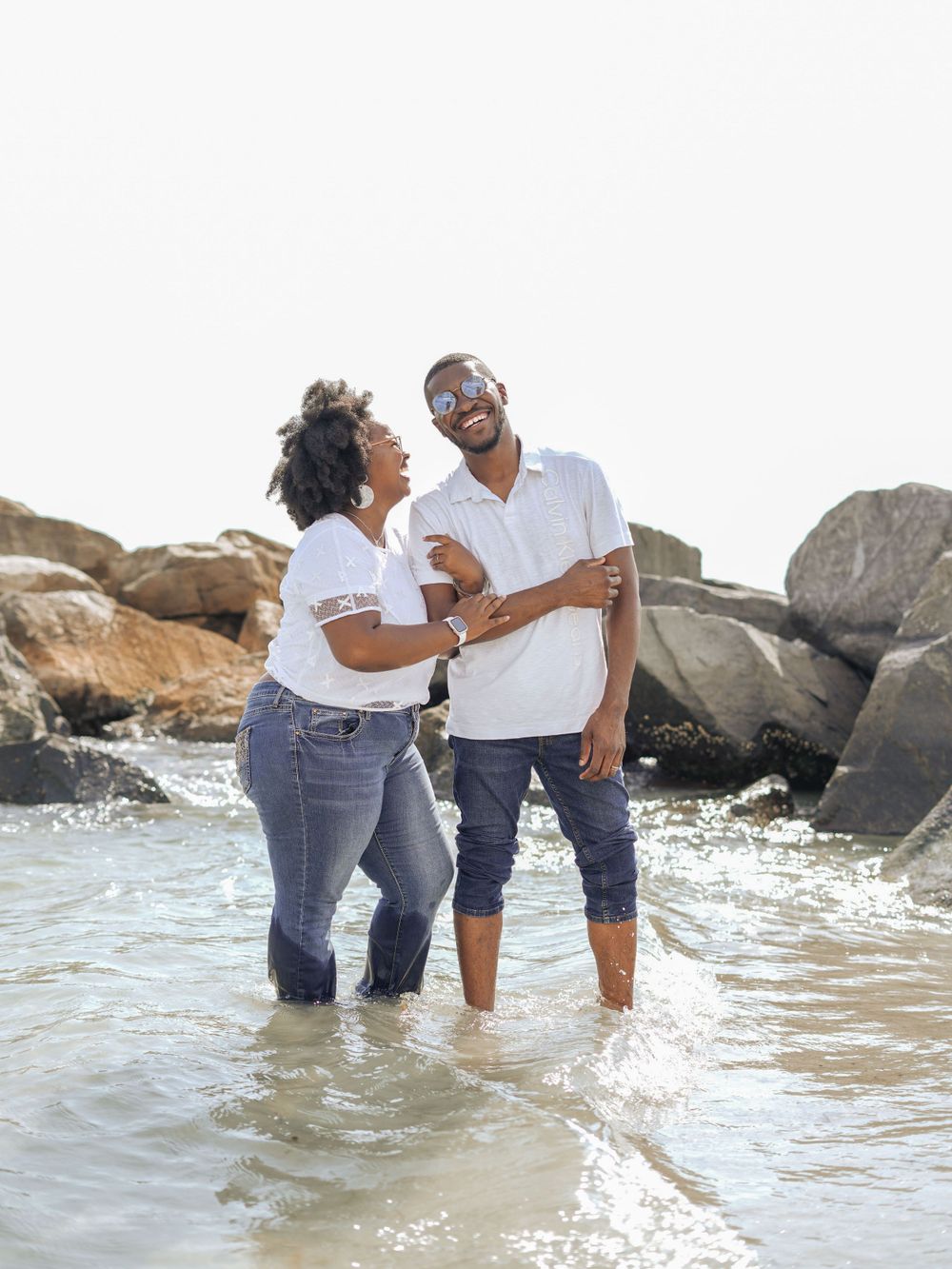 Couple laughs together while standing in shallow ocean water near rocky shore.