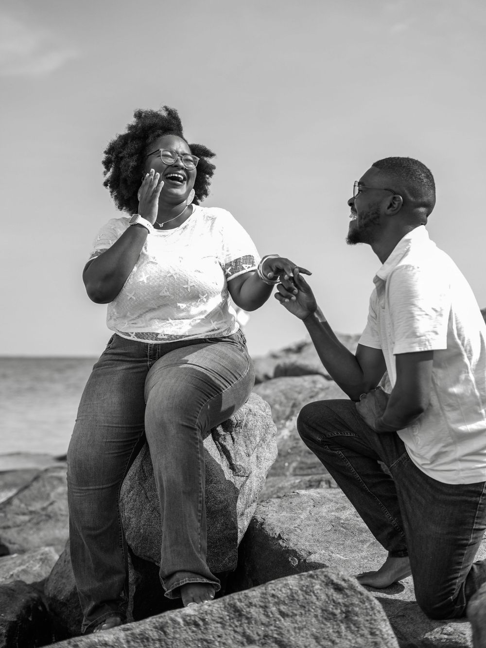 Black and white photo captures beach proposal moment on rocky shoreline.