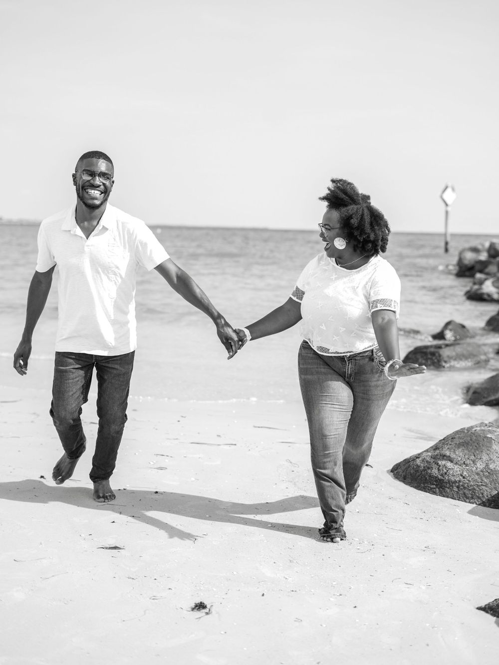Black and white photo of couple walking hand in hand along beach with rocks.