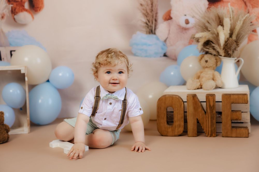First birthday party setup with wooden 'ONE' letters, blue balloons, and floral decorations against beige backdrop.
