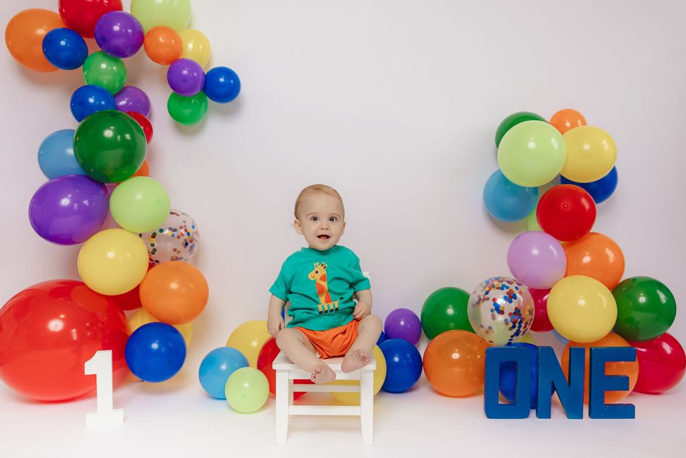 Colorful birthday balloon garland decorates a white backdrop with blue letter blocks spelling 'ONE' and a small white chair.