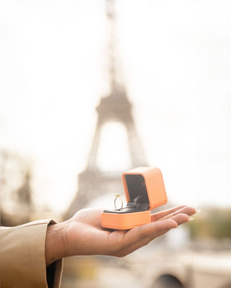 An engagement ring box held in front of the blurred silhouette of the Eiffel Tower.