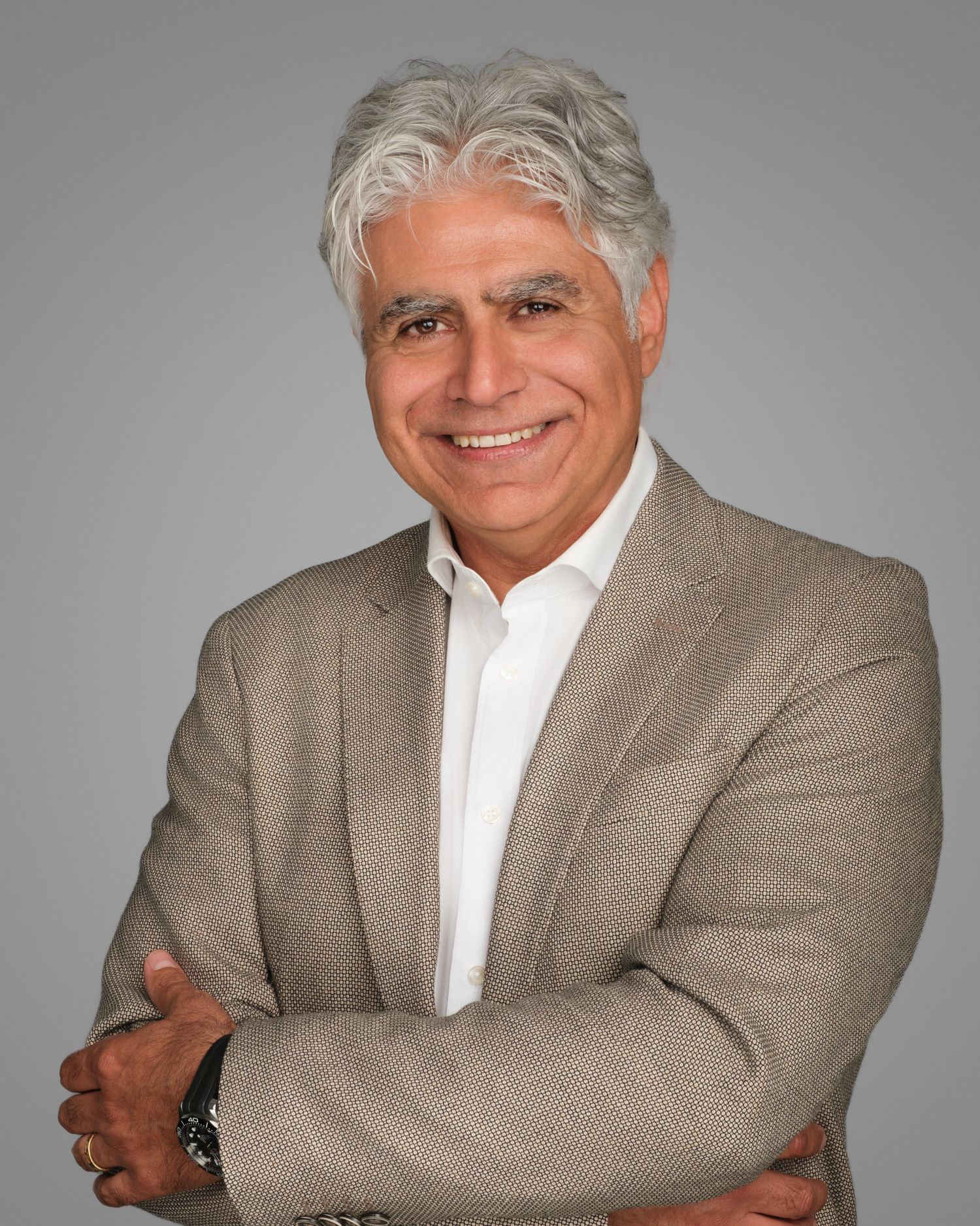 A professional headshot of a person with silver hair wearing a beige suit jacket and white shirt against a gray background.