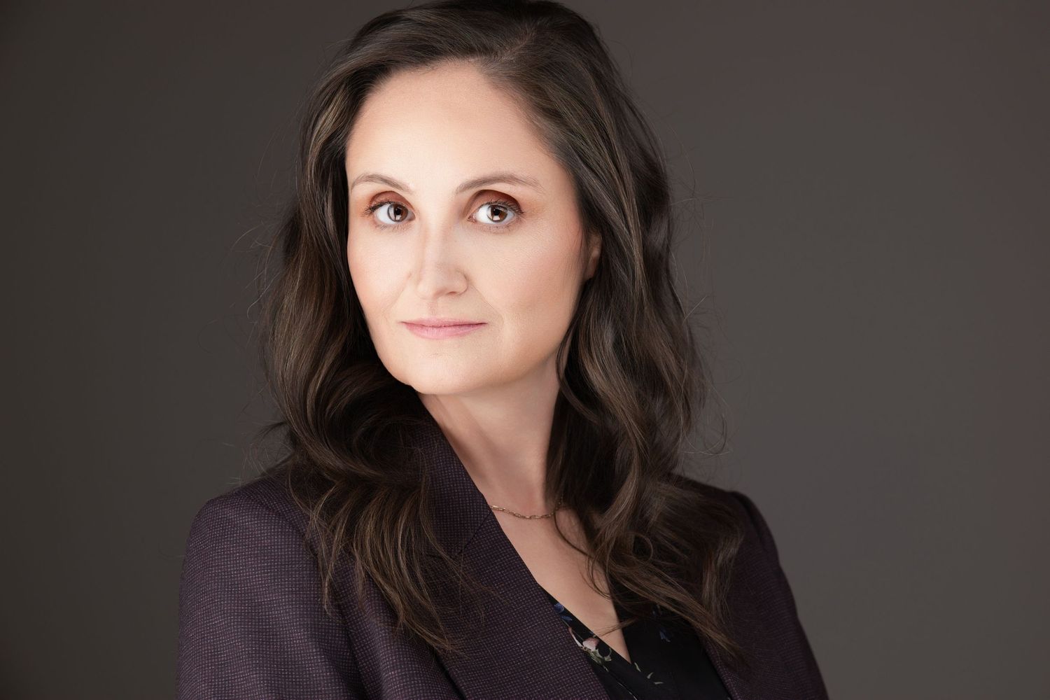 Professional studio portrait of someone with long dark hair wearing a dark blazer against a gray background.