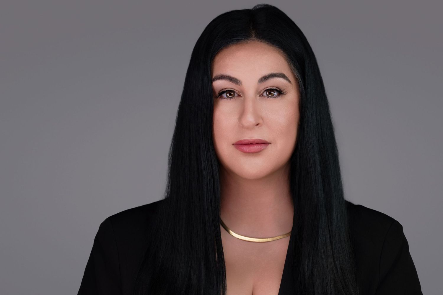 Professional studio portrait of a person with long black hair wearing a black blazer and delicate necklace against gray background.