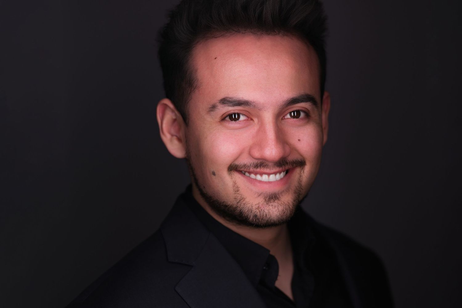 Professional headshot portrait of smiling person wearing dark blazer against black background with dramatic lighting.