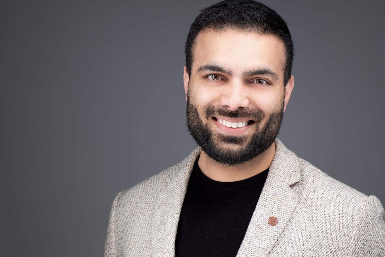 Professional headshot portrait against gray background of a smiling person wearing a beige blazer and black shirt.