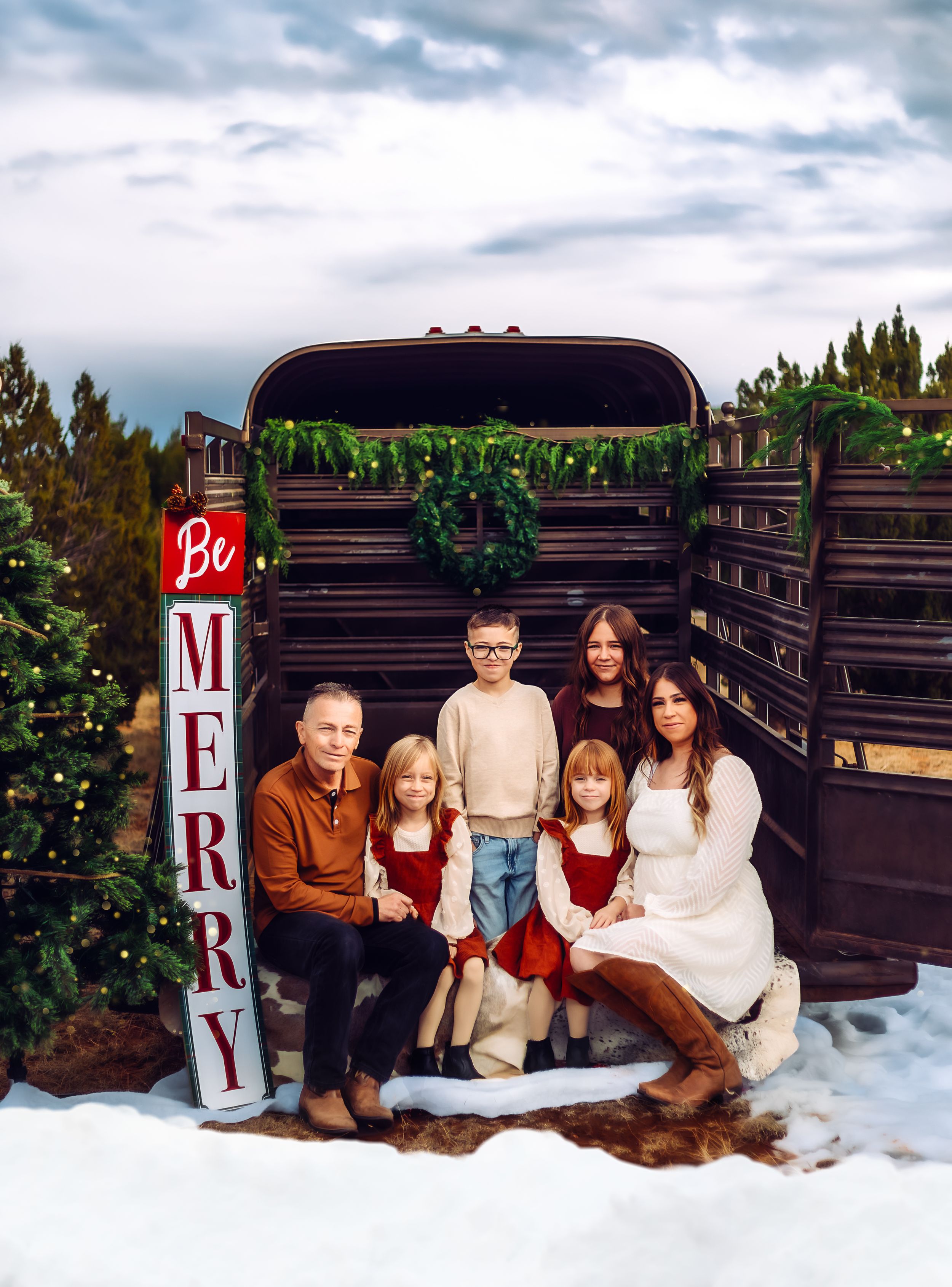 A family poses on wooden benches in a winter-themed photoshoot setup with snowy backdrop and festive holiday decor.
