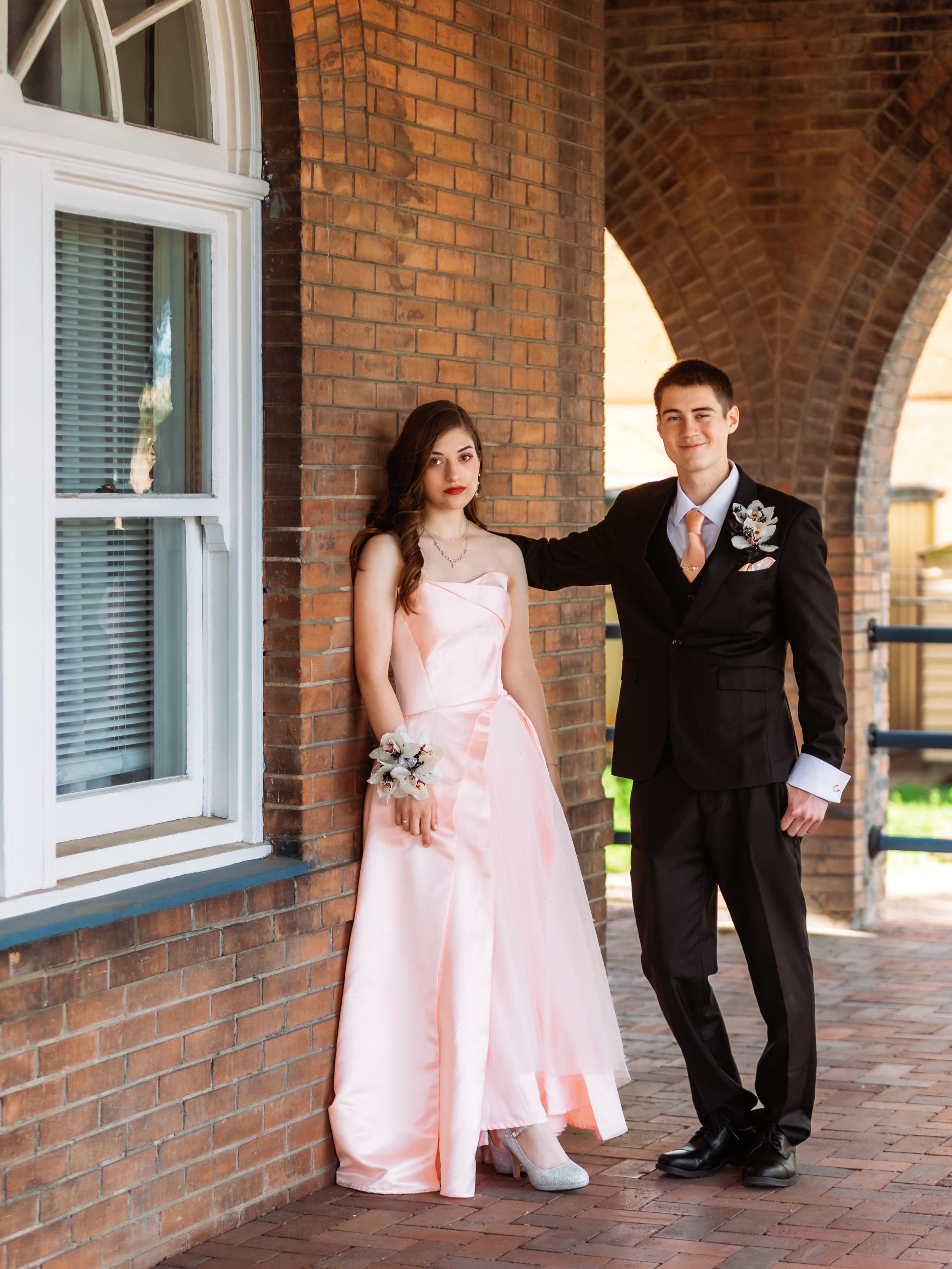 Two children in formal attire - one in pink dress, one in black suit - stand against a brick wall in candid poses.