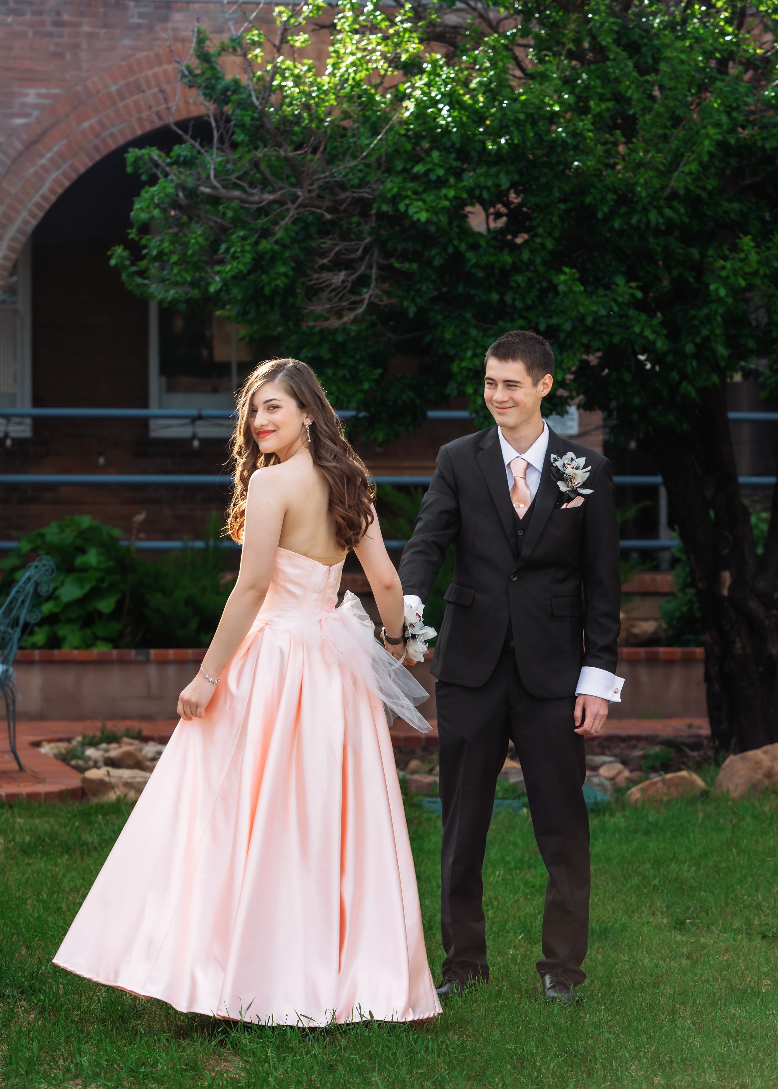 A couple in formal attire poses for romantic photos in a garden with brick buildings in the background.