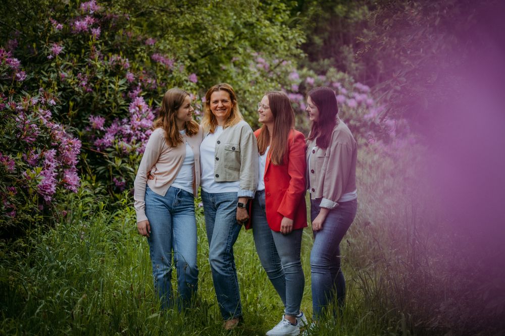 Four friends stand closely together in a lush garden, smiling amidst vibrant pink rhododendron flowers and green grass.