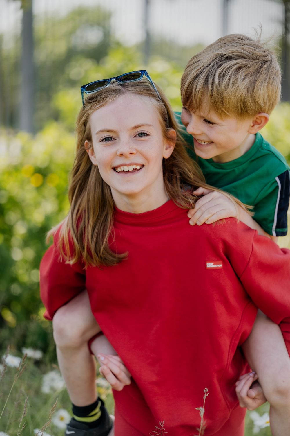 A child in a red sweater gives a piggyback ride to a younger child in a green shirt, surrounded by greenery and wildflowers.
