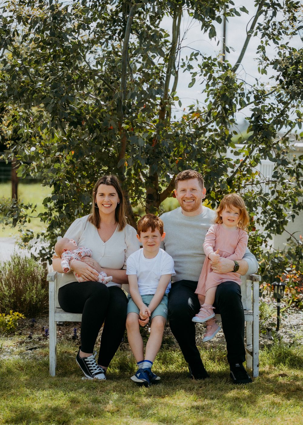 A family of five poses together on a rustic bench surrounded by lush greenery in a sunny outdoor setting.