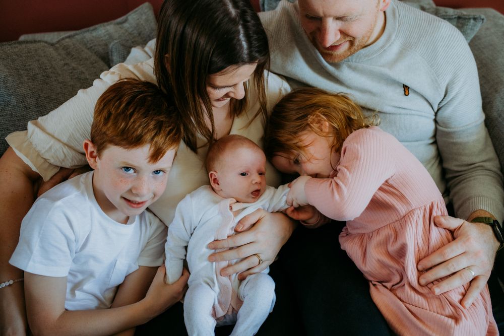 A family of four sits closely on a couch, with two children cuddled and a baby held lovingly in the mother's arms.