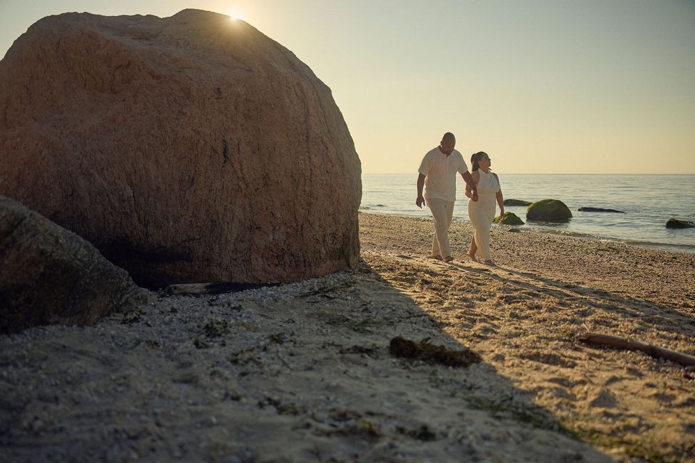 A couple walking along a beach at sunset with large rocks and boulders silhouetted against the golden sky.